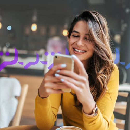 Photoillustration of a woman holding an iPhone in both of her hands and smiling at the screen; she has long, wavy brown hair and light coffee-colored skin and she's wearing coral lipstick; on top of the photo, there's an overlay graphic of message bubbles